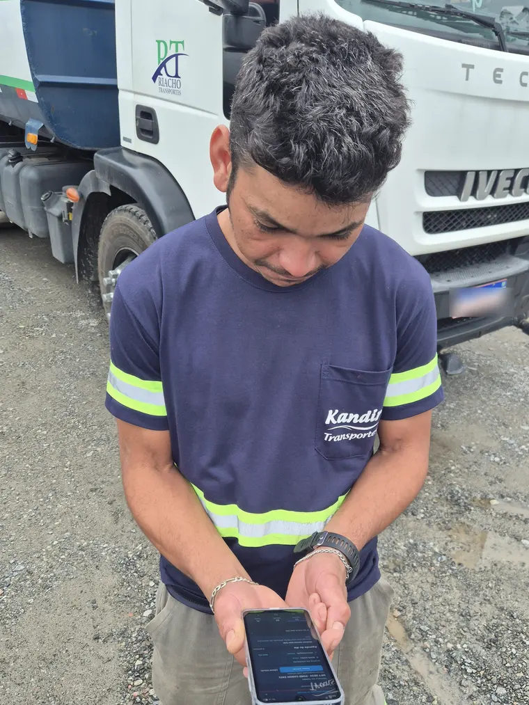 Motorista no pátio, ao lado do caminhão, consulta a agenda do dia no celular.